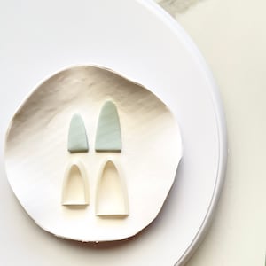 Pode incluir: Dois pares de brincos de argila polimérica, um par é de cor azul claro e o outro par é um molde branco. Os brincos têm a forma de um arco.