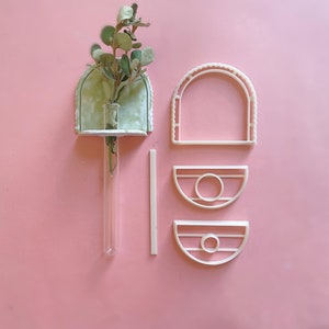 May include: A set of white ceramic wall planters in the shape of arches and half circles. The planters are designed to hold small plants or flowers. A small glass vase with a sprig of greenery is placed in the arch planter.