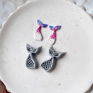 May include: Four mermaid tail charms, two are gray and two are pink, blue, and white.