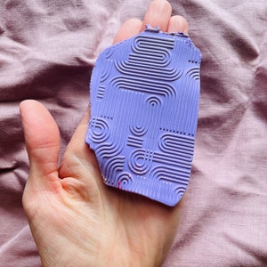 May include: A purple rubber stamp with a geometric pattern. The stamp is slightly chipped on the top edge.