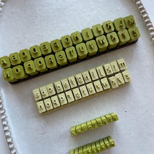 Pode incluir: Vista aérea de carimbos de letras em diferentes tamanhos e cores numa bandeja oval branca. Os carimbos são em fonte de bloco, com tons de verde e bege. Ideal para artesanato e personalização.