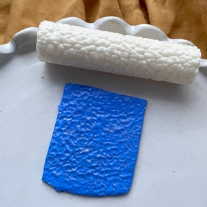 Op de afbeelding: Een witte textuurroller en een blauwe textuurplaat van polymer clay. De roller wordt gebruikt om textuur op de klei te creëren.