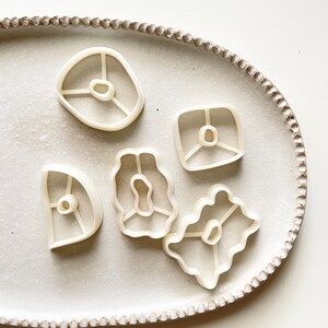 May include: A set of six white plastic cookie cutters in various shapes, including a square, a circle, and a few organic shapes. The cutters are arranged on a white oval plate with a textured rim.