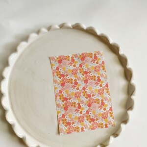 May include: A white ceramic plate with a scalloped edge, featuring a pink and orange floral patterned paper card in the center.