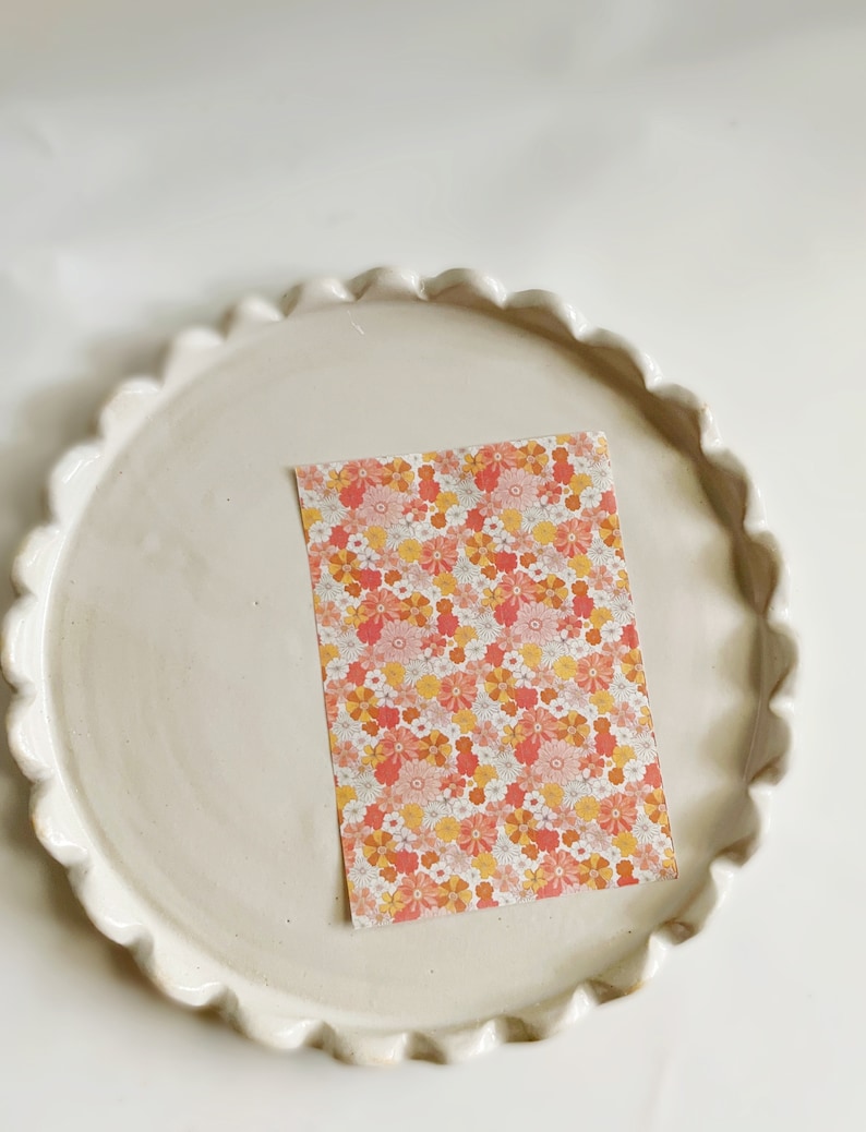 May include: A white ceramic plate with a scalloped edge, featuring a pink and orange floral patterned paper card in the center.