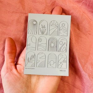 Könnte beinhalten: Eine Bogen mit 12 Nagelaufklebern mit verschiedenen Bogen-Designs, die Naturmotive wie Sonne, Berge, Wolken und Pflanzen zeigen. Die Aufkleber sind auf einem hellgrauen Hintergrund gedruckt und haben den Text "Phoenix & Spruce" und "10CT-11" unten aufgedruckt.