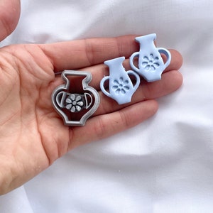 May include: Two light blue ceramic vase cookie cutters and a silver metal vase cookie cutter. The ceramic cutters have a flower design on the front.