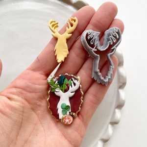 May include: Three reindeer ornaments, one is a white reindeer with green holly and red berries, one is a yellow reindeer, and one is a gray reindeer cookie cutter.