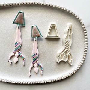 May include: Two pink and white polymer clay earrings shaped like a gymnast doing a handstand. The earrings have a teal and purple striped design. The earrings are on a white plate with two white polymer clay molds.