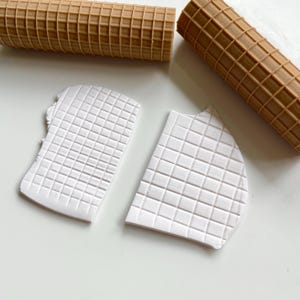 May include: Two brown textured rolling pins and two white clay pieces with a grid pattern. The rolling pins are cylindrical with a waffle-like design. The clay pieces have a similar grid pattern pressed into them.