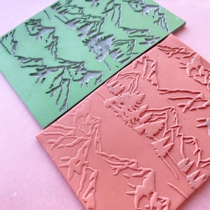 May include: Two rubber stamps with mountain and tree designs. The top stamp is green and the bottom stamp is orange.