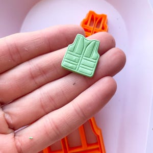 May include: A green plastic mold of a life vest. The mold is being held in a hand. The mold is used to create fondant or gum paste decorations for cakes.