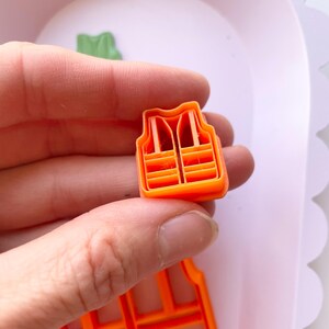 May include: Orange plastic cookie cutter shaped like a life vest. The cutter has a detailed design with multiple horizontal lines.