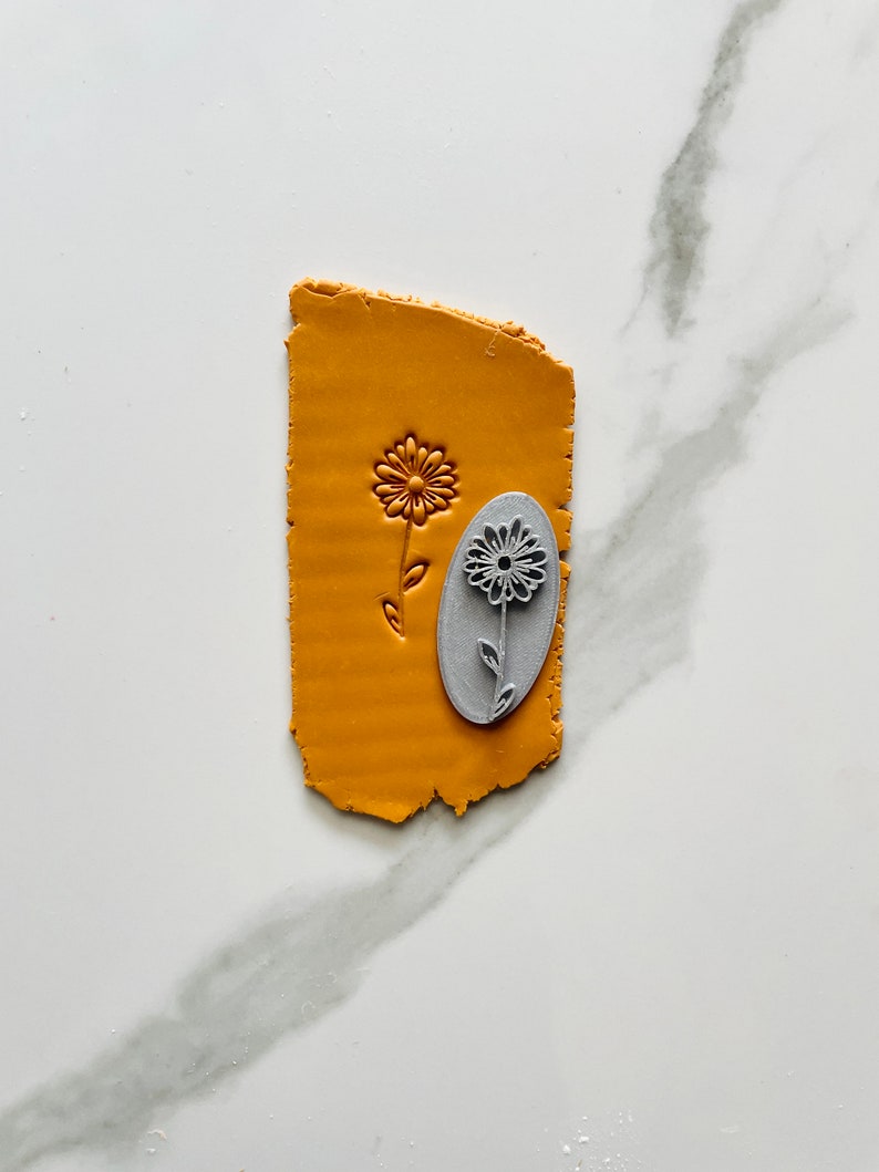 Photo of a clay stamp for polymer clay representing a zinnia flower, looking similar to a daisy. Ian the photo is also a sample of mustard clay textured with the same stamp.