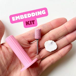 Puede incluir: Un kit de incrustación rosa con una broca de lijado cilíndrica, una herramienta rosa en forma de columna y un objeto blanco en forma de concha. El kit está diseñado para manualidades e incluye el texto "EMBEDDING KIT" en una pancarta rosa.