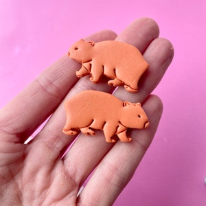 May include: Two orange wombat-shaped embellishments. The wombat figures are a solid color with visible details like ears, legs, and a small tail. The background is a solid pink color.