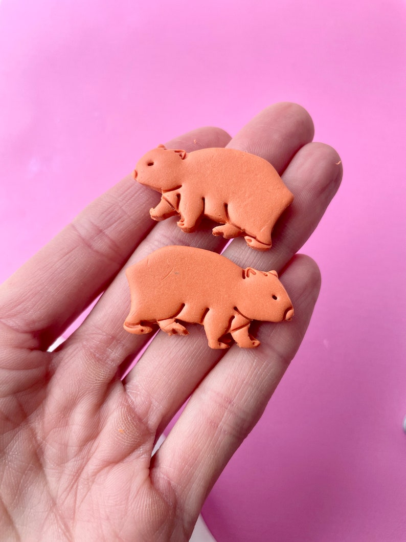 May include: Two orange wombat-shaped embellishments. The wombat figures are a solid color with visible details like ears, legs, and a small tail. The background is a solid pink color.