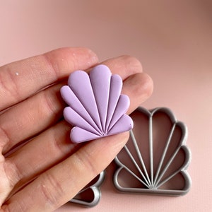 Photo of a lilac Art Deco polymer clay earring held by a hand. In the background is the clay cutter that has been used to make this earring. The shape resembles a seashell.