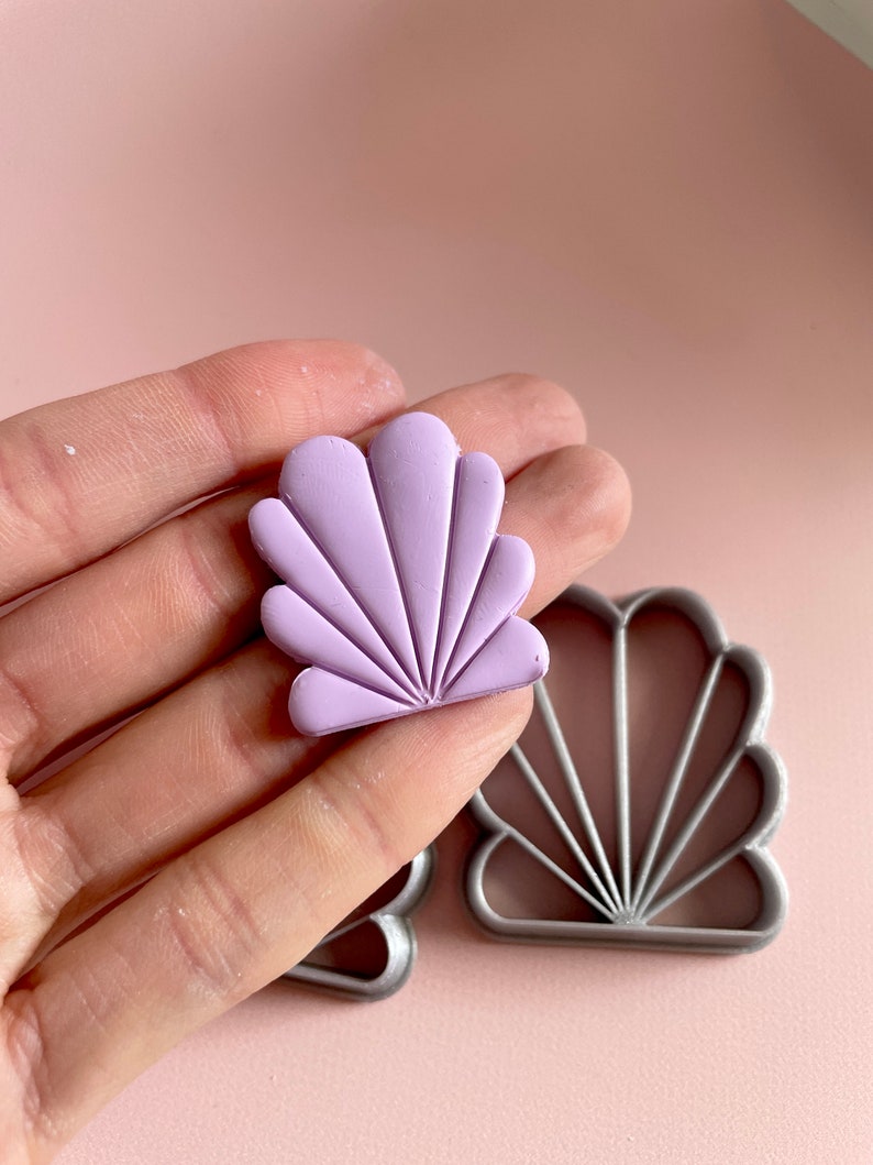 Photo of a lilac Art Deco polymer clay earring held by a hand. In the background is the clay cutter that has been used to make this earring. The shape resembles a seashell.