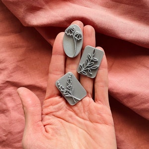 May include: Three grey rubber stamps with floral designs. The stamps are in the shape of an oval and two rectangles. The designs are of a flower, a branch with leaves, and a sprig of lavender.