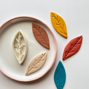 May include: Five leaf-shaped cookie cutters, two are brown, one is beige, one is blue, and one is yellow. The cutters are on a white surface.