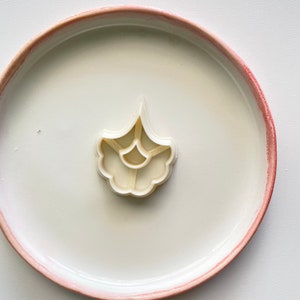 May include: White plastic cookie cutter shaped like a flower with five petals. The cutter is on a white plate with a pink rim.