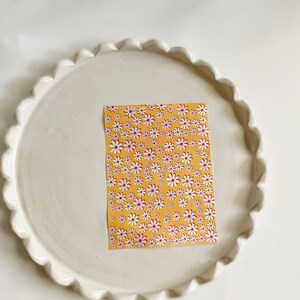 May include: A white ceramic plate with a scalloped edge, featuring a yellow sticker with a repeating pattern of pink and white daisies.