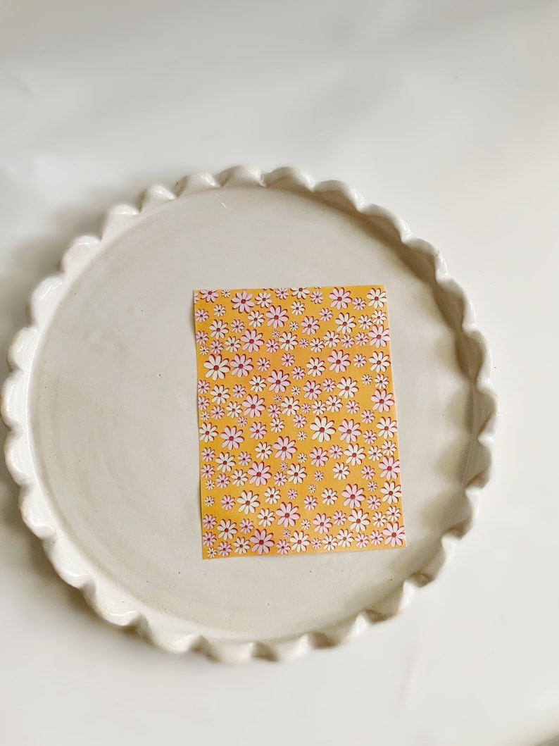 May include: A white ceramic plate with a scalloped edge, featuring a yellow sticker with a repeating pattern of pink and white daisies.