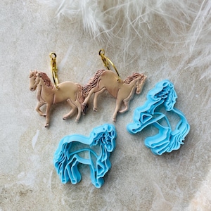 Puede incluir: Un par de pendientes de aro dorados con un dije de caballo marrón y un par de dijes de caballo azules. Los dijes están hechos de arcilla polimérica y tienen forma de caballos.