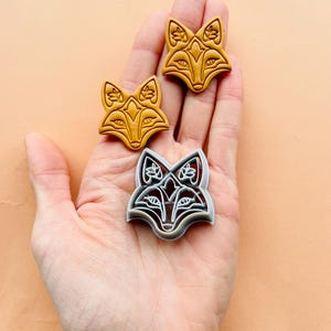 Puede incluir: Tres cortadores de galletas de zorro, dos son dorados y uno es plateado. Los cortadores están detallados con diseños florales y tienen un patrón geométrico.