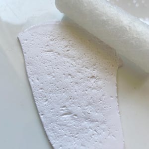 May include: A close-up shot of a white, textured clay slab with a white rolling pin. The clay has a rough, porous surface. The rolling pin is cylindrical and also white. The items are on a white surface.