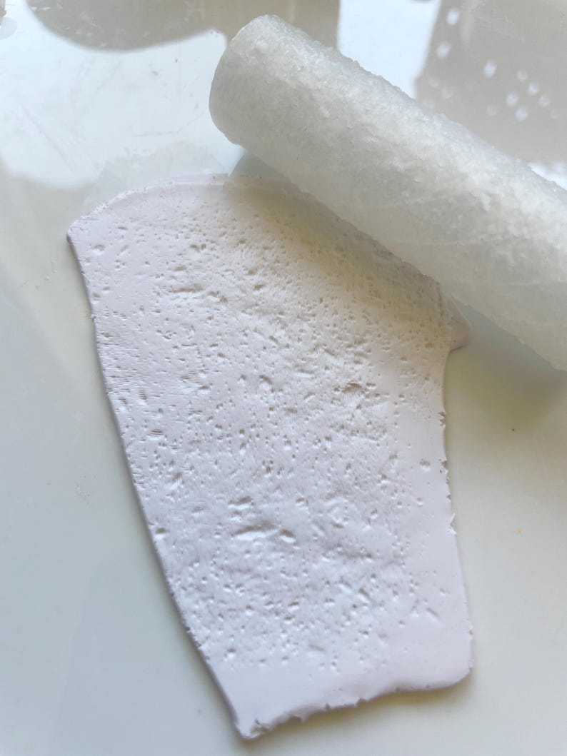May include: A close-up shot of a white, textured clay slab with a white rolling pin. The clay has a rough, porous surface. The rolling pin is cylindrical and also white. The items are on a white surface.
