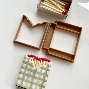 Puede incluir: Un juego de dos cortadores de galletas de plástico marrón, uno con forma de caja de cerillas y otro con forma de cerilla, junto a una pequeña caja de cartón llena de cerillas de madera con puntas rojas y una galleta con forma de caja de cerillas y patrón de cuadros verdes.
