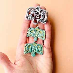 May include: Two teal blue ceramic bird ornaments with gold accents. The ornaments are in the shape of a stylized bird with outstretched wings. The bird has a floral design on its wings and chest. A gray plastic mold of the same bird design is also shown.