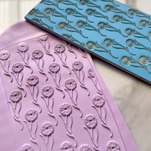 Puede incluir: Dos sellos de silicona con un diseño de diente de león. El sello superior es azul claro y el sello inferior es morado claro.