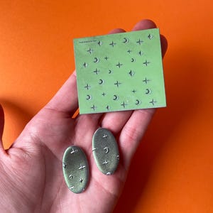 Può includere: Un paio di orecchini ovali verdi con un design a forma di luna e stella. Gli orecchini sono mostrati su una mano che tiene un foglio di carta verde con un motivo di lune e stelle.