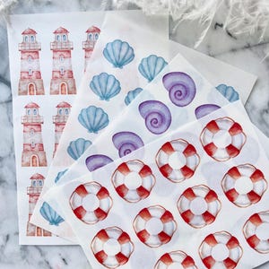 May include: Sheets of watercolour stickers with nautical themes. Designs include red and white lighthouses, blue seashells, purple spiral shells, and red and white life preservers. The stickers are arranged on white paper.