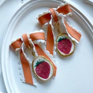 Pu&ograve; includere: Orecchini fatti a mano con motivo a fragola. Ogni orecchino ha una cornice color oro attorno a un'immagine di fragola rossa, sospesa da un fiocco con nastro arancione e finiture smerlate bianche. Gli orecchini sono esposti su una superficie bianca.