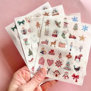 Könnte beinhalten: Eine Hand hält mehrere Bögen mit Aufklebern zum Thema Weihnachten. Die Aufkleber zeigen festliche Illustrationen wie Lebkuchenmänner, Schneekugeln, Weihnachtsbäume und Rentiere in verschiedenen Farben, darunter Rot, Grün und Gold. Der Hintergrund ist einfarbig rosa.