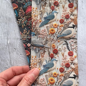 Puede incluir: Dos hojas de papel decorativas con diseños en relieve. Una presenta un fondo oscuro con setas y follaje. La otra muestra pájaros azules, flores y hierbas en un campo blanco, naranja y rosa.