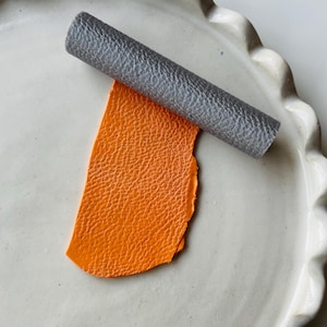 Peut inclure: Gros plan sur un morceau de cuir orange vif en cours de roulage avec un rouleau texturé gris sur une assiette en céramique blanche.