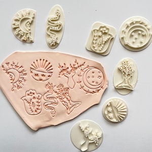Puede incluir: Un conjunto de nueve sellos de goma blanca con varios diseños, que incluyen una luna creciente, un sol, una serpiente, una mano, un árbol y una flor. Los sellos están dispuestos sobre una superficie rosa claro.