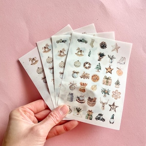 May include: A hand holds several sheets of Christmas-themed stickers. The stickers feature watercolor illustrations of ornaments, rocking horses, trucks, stars, and other holiday elements. The stickers are on a pink background.