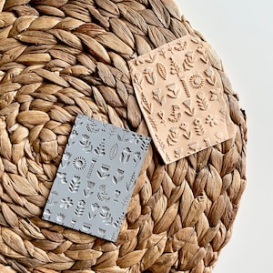 May include: Two rectangular rubber stamps with floral designs. The stamps are on a woven brown surface. One stamp is gray and the other is light brown.