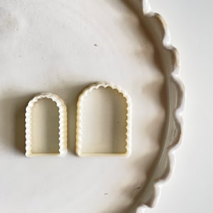 May include: Two white plastic cookie cutters shaped like arches with scalloped edges. The cutters are on a white surface.