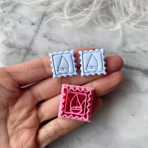 Puede incluir: Tres cortadores de galletas con forma de sello. Dos son de color azul claro y uno rosa. Cada cortador presenta un diseño de velero dentro de un marco rectangular con un borde festoneado. Los cortadores probablemente se utilizan para hornear o manualidades.