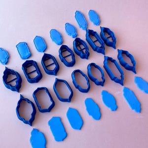 Puede incluir: Un conjunto de 18 cortadores de galletas de plástico azul en varias formas, incluyendo rectángulos, óvalos y diseños ornamentados. Los cortadores están dispuestos en filas sobre una superficie rosa.