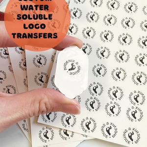 May include: A stack of white stickers with a black circular logo that says "Phoenix & Spruce" with a hand gesture inside the circle. The text "Custom Water Soluble Logo Transfers" is written in black on a red circle in the upper left corner of the image.
