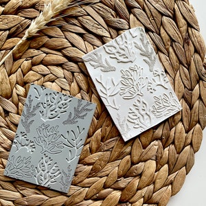 May include: Two rectangular embossed tiles with a coral reef design. One tile is white and the other is grey. The tiles are on a woven straw mat.