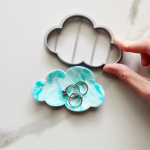 Puede incluir: Un porta anillos en forma de nube de color azul turquesa con vetas blancas. El porta anillos está lleno de cuatro anillos, incluyendo un anillo de plata con una piedra preciosa azul.